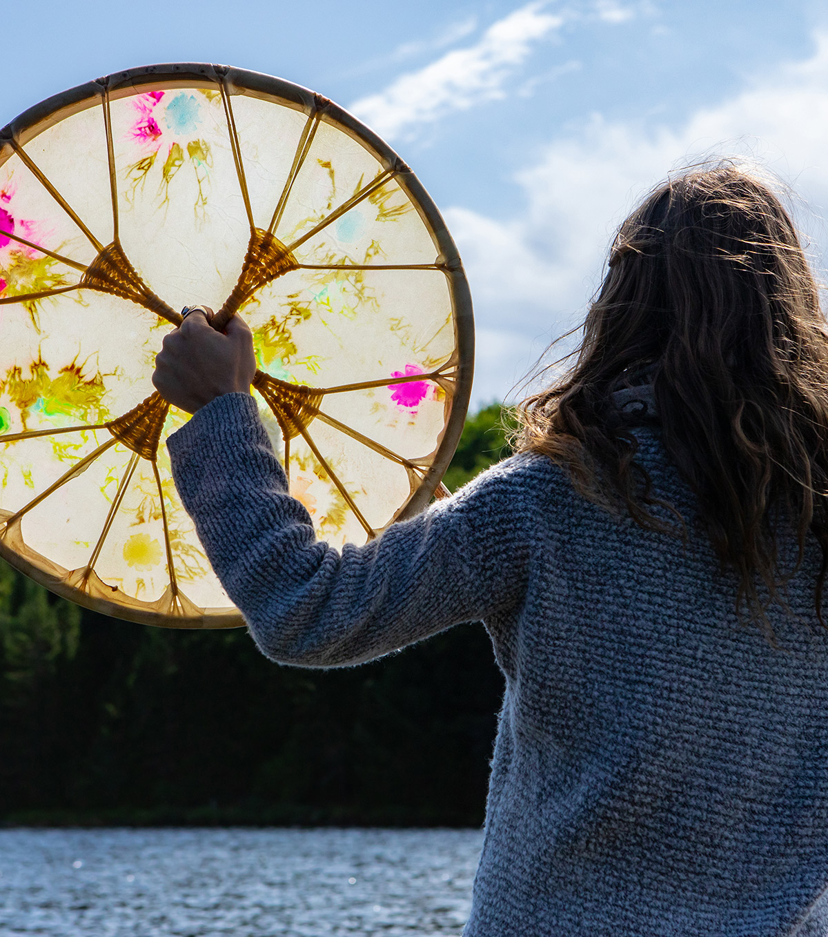 Personne de dos tenant un tambour à cadre sacré autochtone devant un lac 