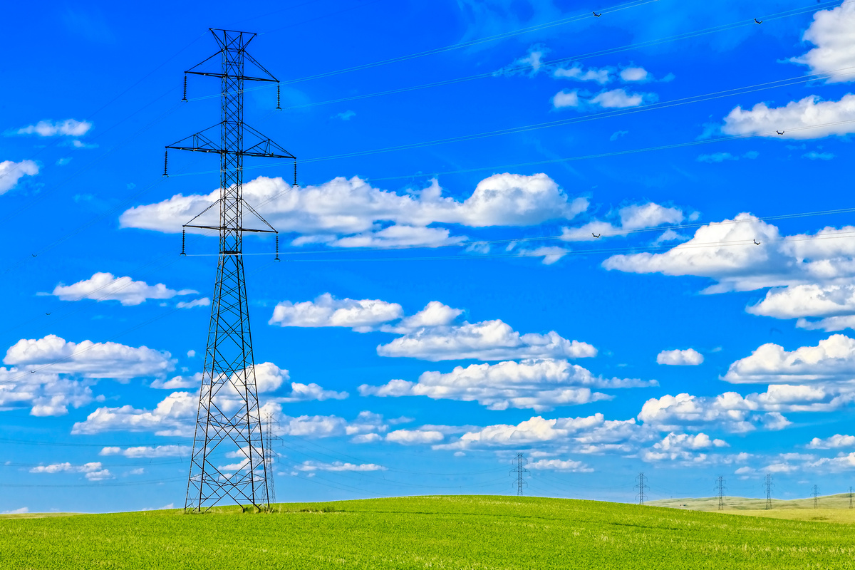 Les pylônes qui relient les lignes de transport d’électricité traversent un champ vert. Le ciel est d'un bleu éclatant, parsemé de nuages blancs cotonneux.