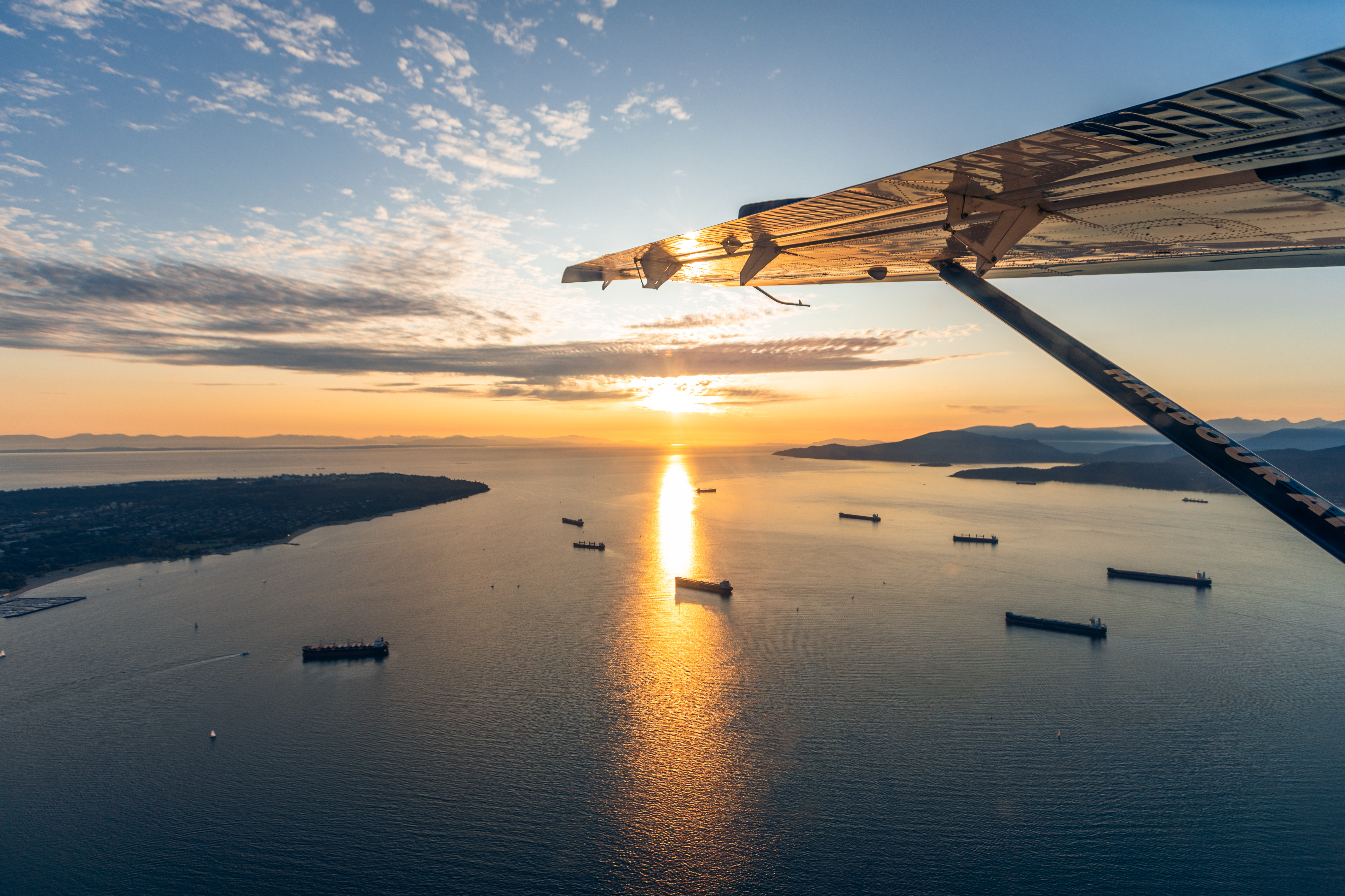 Aile d’un avion de Harbour Air survolant la baie English au coucher de soleil, avec des navires-citernes ancrés en mer en contrebas.