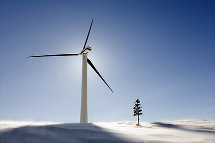 Snowy windy landscape with a wind backlit wind turbine and a lonely spruce tree in the foreground.