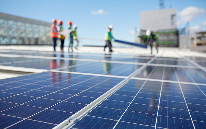 Solar panels in the foreground, construction workers in PPE are unfocused in the background installing solar panels onto a roof.